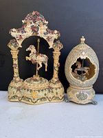 Two decorative horse musical boxes side by side. Left is an ornate floral arch with a carousel horse suspended in the center and gold accent base. Right is an egg-shaped music box with detailed embossing and a carousel horse inside.