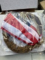 Photo showing two 20 inch grapevine wreaths decorated with red, white, and blue striped fabric and burlap ribbons, packaged in plastic wrapping. Box labeled Orchid & Lily seen behind.