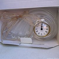 Clock in open box showing front clear glass shell-shaped clock with white face and gold ribbon