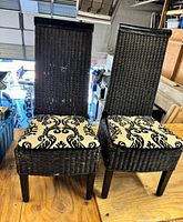 Two black wicker patio chairs with high backs and black legs, each with a cream and black patterned seat cushion. Photo taken from front angle to show chairs' design and condition.