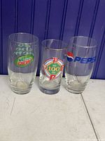 Three clear drinking glasses with logos of Mountain Dew, Detroit Tigers 100th Anniversary, and Pepsi, arranged side by side against a blue wood background and white surface.