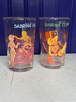 Front view of two clear glasses with colorful comic-style illustrations of Sabrina characters in various scenes.