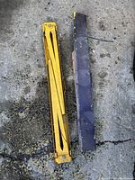 Folded yellow metal sawhorse leg support with visible wear, next to black metal component on a rough concrete surface.
