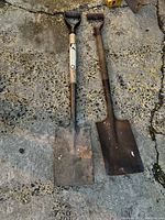 Two used garden shovels side by side on rough concrete ground, showing overall condition and blade shapes.