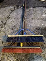 Two push brooms laid on pavement, one with black bristles and a squeegee, the other with red bristles.