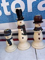 Front view of three snowman pottery candleholders showing details of hats, scarves, and faces.