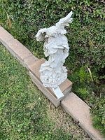 Full view of white angel statue with harp, visible detail of wings, robes, and base, set on outdoor brick edge with hedge background.