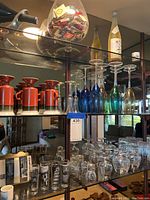 Photo of multiple shelves containing assorted bar glassware including stemmed glasses, ceramic mugs, two bottles, and a large bowl filled with matchboxes.