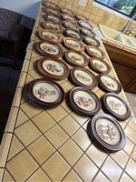 25 framed Hummel plates in a row on a kitchen counter showing oval wooden frames and embossed colorful images of children, marked by years 1971-1995.