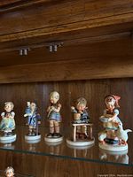 Five Hummel figurines on a glass shelf with wooden background, showing each figurine in full.