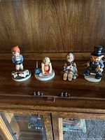 Four Hummel figurines arranged on a wooden shelf showing detailed ceramic figures of children in various poses with one boy with pigs, one girl with umbrella, one seated girl, and one boy in hat.