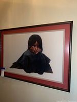 Framed art of a woman with dark hair resting her face on her hands, dark textured top, with red and beige mat and dark brown frame.