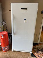 Front view of a white upright freezer with a vertical handle on left, standing on floor.