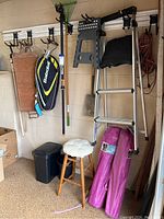Photo showing corner of garage with small step ladder hanging on wall, a wooden stool with white cushion, two pink folded Embark chairs in bags, and black trash cans on floor. Farming and garden tools hanging above.