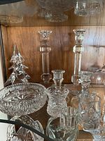Photo showing two tall faceted glass candlesticks, smaller candle holders, cut glass pedestal dish, and glass Christmas tree decoration in a wooden display cabinet.
