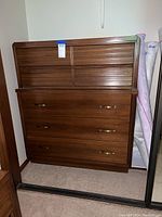 Front view of a medium brown wooden highboy dresser with five drawers, two smaller upper drawers with horizontal slats and three larger lower drawers with brass handles.