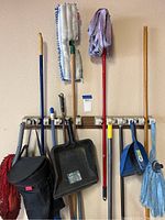 Full view of mops, brooms, dustpans, caddy bag and gloves hanging on wall hooks showing condition and variation of cleaning tools.