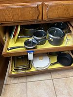 Kitchen cabinet drawer with multiple black pots and pans with glass lids, cast iron skillet, white CorningWare baking dish, Pyrex glass dishes and cutting boards.