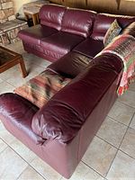 L-shaped leather couch in burgundy with two pillows on the seating area and a colorful throw blanket on the backrest.