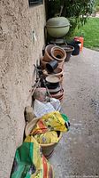 Multiple stacked terracotta and black plastic plant pots with two bags of potting mix in front and a green dome BBQ grill in background.