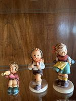 Three Hummel figurines on glass shelf showing child playing trumpet, boy running with book/box, girl in yellow apron with red bow
