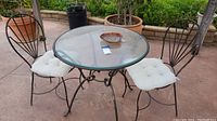 Full view of wrought iron round patio table with glass top and two matching chairs with white cushions on outdoor patio.