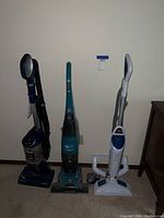 Photo of three floor cleaning devices standing upright against a wall: a dark blue upright vacuum, a turquoise stick vacuum, and a white steam mop.