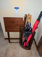Photo showing the umbrella stand containing multiple umbrellas and a pair of black gloves, with the folding table behind it against the wall.