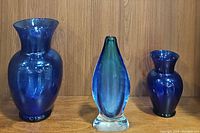 Front view of the three blue vases displayed side by side on a wooden shelf: large cobalt vase, teardrop glass vase with clear base, and smaller cobalt vase.