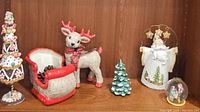Vintage Christmas ceramic and wooden figurines arranged on a shelf including a gumdrop tree, reindeer, sleigh, ceramic tree, Santa Claus figure, and globe ornament.