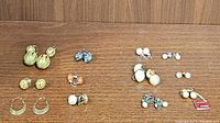 Top view of assorted stud and clip-on earrings arranged on a wooden surface, showing various pairs in gold-tone, silver-tone, pearl-like and colored styles.