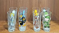 Front view of seven clear glass tumblers arranged in two rows, each with different Looney Tunes character decals including Bugs Bunny, Daffy Duck, Tweety Bird, Road Runner, Wile E. Coyote, Elmer Fudd, and Yosemite Sam.