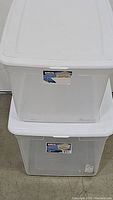 Two clear plastic storage totes stacked, each with white lids and labels visible on the tactile side, showing slight wear and use.