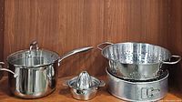 Photo of stainless steel saucepan with glass lid, a springform pan, a colander, a metal hand juicer, and two quiche pans stacked inside the colander.