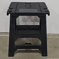 Front view of the Stanley foldable plastic workbench showing the open legs, a black perforated top surface, and a rectangular inset compartment with Stanley logo.