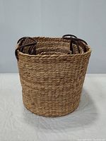 Single largest basket shown standing upright on quilted surface, highlighting texture and leather handle loops attached to basket sides.