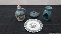 Four items arranged on a black cloth: Emerson Creek soap dispenser, blue ceramic oil burner, Whieldon Ware floral saucer plate, and a small blue teapot-shaped item.