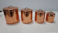 Front view of four copper canisters with brass lid handles arranged largest to smallest