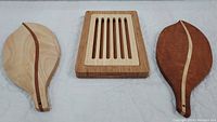 Three cutting boards pictured flat on white surface, showing rectangular bread board with vertical slots in center and two leaf-shaped cutting boards with wood inlays on each side