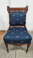 Full frontal view of the Victorian chair showing blue fabric upholstery with embroidered patterns, dark wood frame, and turned legs on original wooden casters.