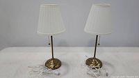 Two brass-tone IKEA Arstid table lamps with white pleated fabric shades, showing full lamps with cords on a white backdrop.
