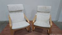 Photo of two IKEA POANG armchairs side by side showing birch wood frames and white cushioned seats and backs.