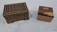 Two wooden boxes with lids. One is rectangular with detailed floral inlay on all visible surfaces. Second is a small square cube with a lid showing a mountain scene inlays in various wood tones.