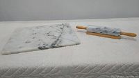 Overall view of marble cutting board on left and marble rolling pin with wooden handles resting on wooden holder on right on a white quilted surface.