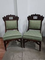 Front view of two matching Eastlake style carved walnut chairs with green upholstered seats and backs. Displayed side by side on wood floor.