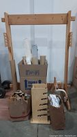 Complete wooden Navajo loom frame standing upright with cardboard box in the center containing rolled materials and accessories, two bags containing various tools and materials at base.