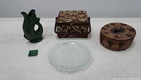Showing the five lot items arranged in a row on white surface: green ceramic fish pitcher, fabric basket caddy, round woven basket, clear flower frog, birch plate.