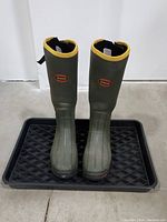 Front view of LaCrosse green rubber boots with yellow rim, placed on a black plastic shoe tray.