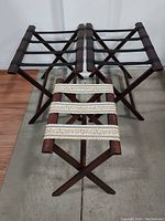 Three vintage wooden luggage racks, two with black straps and one with decorative white/gold straps, set on concrete floor.