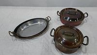 Set of 2 copper pots with lids and an oval copper casserole tray displayed on white quilted surface.